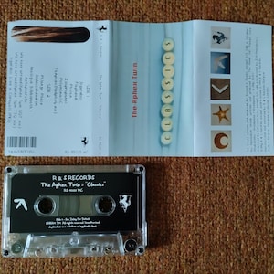 May include: A clear cassette tape with a black label. The label has the text "R & S RECORDS" and "The Aphex Twin - "Classics"" in white. The cassette tape is on a brown surface.