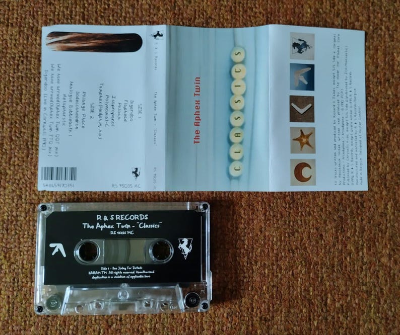 May include: A clear cassette tape with a black label. The label has the text "R & S RECORDS" and "The Aphex Twin - "Classics"" in white. The cassette tape is on a brown surface.