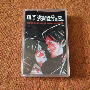 May include: A cassette tape case featuring a black and white image of a couple with red paint splatters on their faces. The text "My Chemical Romance" and "Three Cheers for Sweet Revenge" is printed on the case.