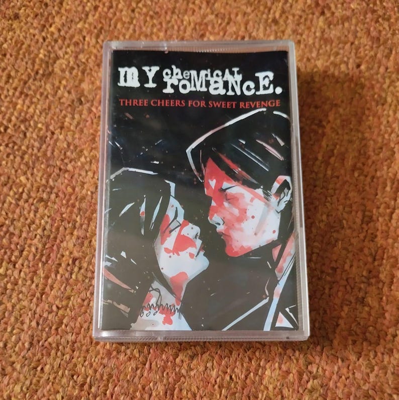 May include: A cassette tape case featuring a black and white image of a couple with red paint splatters on their faces. The text "My Chemical Romance" and "Three Cheers for Sweet Revenge" is printed on the case.