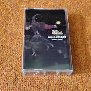 Puede incluir: Una cinta de cassette transparente con una ilustración de una figura, una flor de loto y el texto "TOBIAS FORGE" y "PASSIFLORA" sobre un fondo negro. La cinta está en una caja de plástico transparente.