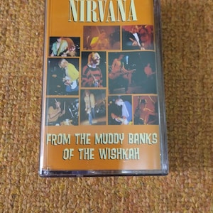 Op de afbeelding: Een cassetteband met een gele achtergrond en de bandnaam "NIRVANA" in zwarte tekst. De band bevat een collage van foto's van de bandleden. De tekst "FROM THE MUDDY BANKS OF THE WISHKAH" is in zwart gedrukt op de onderkant van de band.