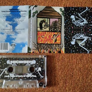 May include: A cassette tape and album art for "Long Term Effects Of Suffering" by Suicideboys. The cassette is clear with a black and white design. The album art features illustrations of skeletons and a house on fire.