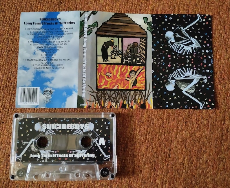 May include: A cassette tape and album art for "Long Term Effects Of Suffering" by Suicideboys. The cassette is clear with a black and white design. The album art features illustrations of skeletons and a house on fire.