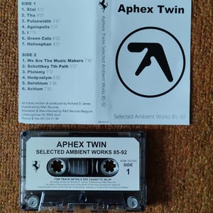 May include: A black cassette tape with a white label that reads "Aphex Twin Selected Ambient Works 85-92". The cassette tape is labeled "Side 1" and has a red Ferrari logo on the left side of the label.