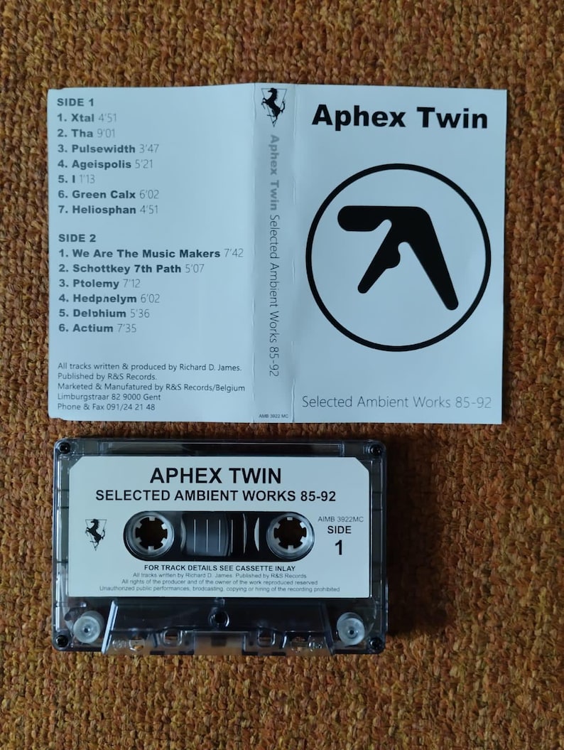 May include: A black cassette tape with a white label that reads "Aphex Twin Selected Ambient Works 85-92". The cassette tape is labeled "Side 1" and has a red Ferrari logo on the left side of the label.