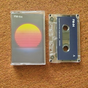 Può includere: Una musicassetta trasparente con un design a tramonto. L'etichetta della musicassetta riporta "FM-84" e presenta un'estetica retrò. La musicassetta è su una superficie marrone.
