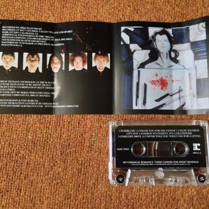 May include: A clear cassette tape with a black label. The label has the text "My Chemical Romance Three Cheers for Sweet Revenge" and the band's logo. The cassette tape is on a brown surface.