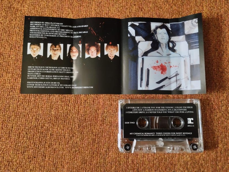 May include: A clear cassette tape with a black label. The label has the text "My Chemical Romance Three Cheers for Sweet Revenge" and the band's logo. The cassette tape is on a brown surface.