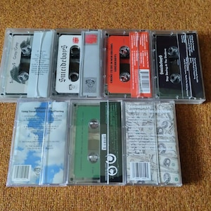 May include: A collection of cassette tapes in clear plastic cases. The tapes feature various colors, including gray, white, red, and black. The text "Suicideboys" is visible on several tapes, along with album titles and track listings.