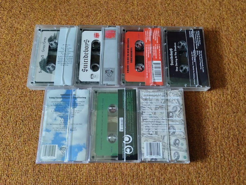 May include: A collection of cassette tapes in clear plastic cases. The tapes feature various colors, including gray, white, red, and black. The text "Suicideboys" is visible on several tapes, along with album titles and track listings.