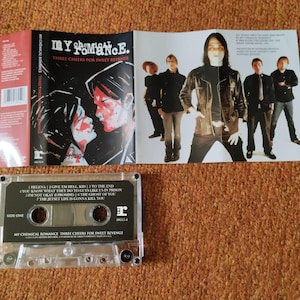 May include: A cassette tape with a clear plastic case and a black label. The label has the band name "My Chemical Romance" and the album title "Three Cheers for Sweet Revenge". The cassette tape is a physical copy of the album.