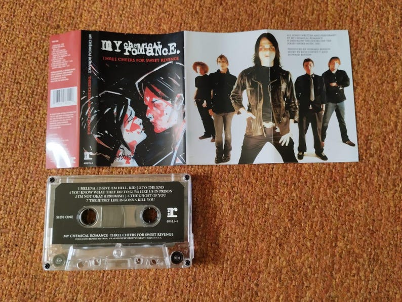 May include: A cassette tape with a clear plastic case and a black label. The label has the band name "My Chemical Romance" and the album title "Three Cheers for Sweet Revenge". The cassette tape is a physical copy of the album.
