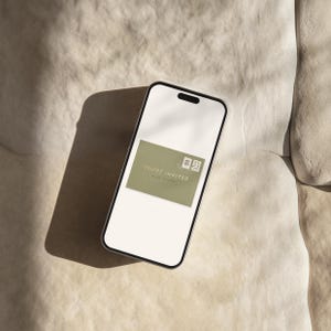 May include: A smartphone displays a digital invitation. The invitation is a sage green rectangle with the words "YOU'RE INVITED" and two small postage stamp graphics. The phone is on a textured, off-white surface.