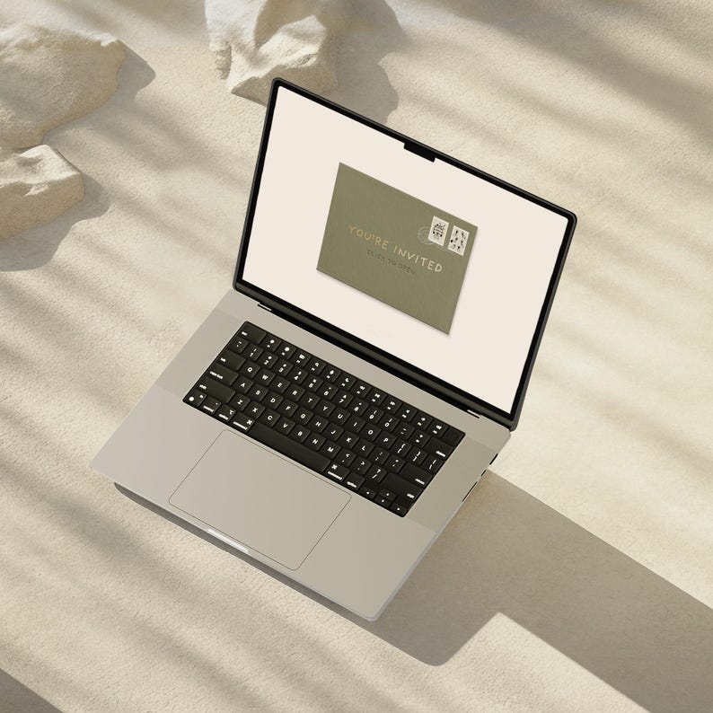 May include: A silver laptop displays a digital invitation on its screen. The invitation is a muted green color with the words "YOU'RE INVITED" and two small postage stamps. The laptop is on a light-colored surface.