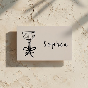 Puede incluir: Una tarjeta blanca con un dibujo de línea negra de una copa con un lazo en el tallo. El nombre "Sophia" está escrito en cursiva negra en la tarjeta.