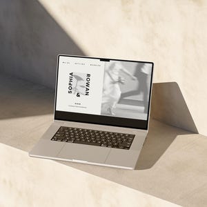 Könnte beinhalten: Ein silberner Laptop zeigt eine Hochzeitseinladung mit den Namen "SOPHIA & ROWAN". Der Bildschirm zeigt ein verschwommenes Bild eines Paares. Der Laptop steht auf einer hellen Oberfläche.