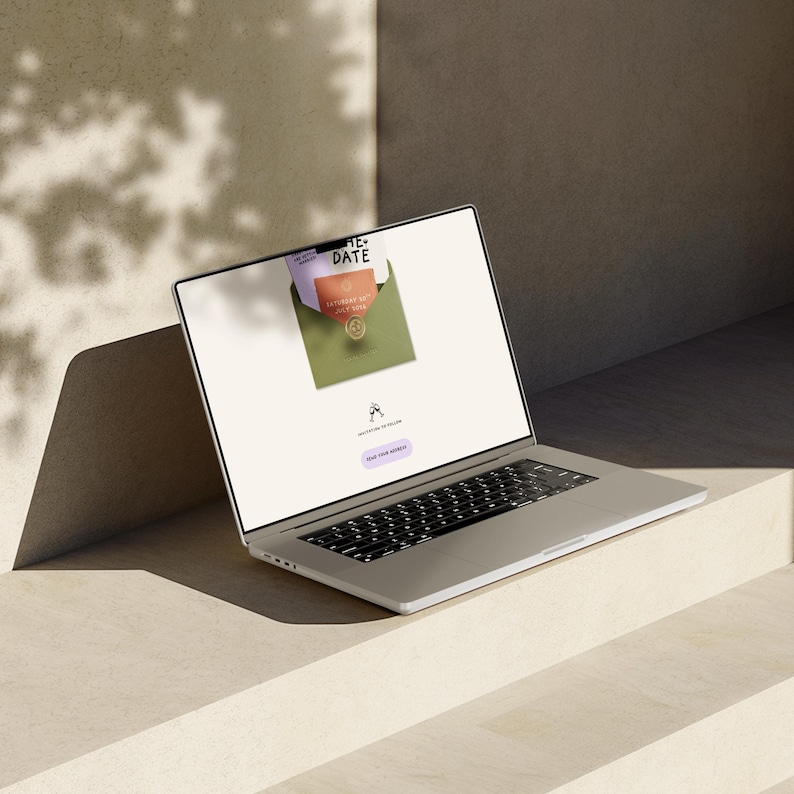 May include: A silver laptop displaying a wedding invitation design. The screen shows an open envelope with invitation cards. The laptop is on a light-colored surface, with shadows from overhead.