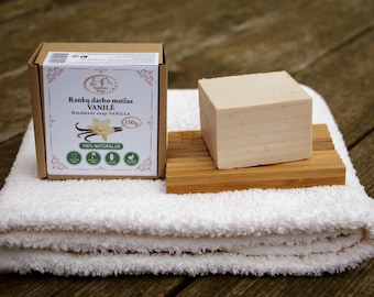 May include: A bar of handmade vanilla soap on a wooden soap dish, with a brown box labeled "Ranku darbo muilas VANILE Handmade soap VANILLA 150g 100% NATURALUS" in the background.