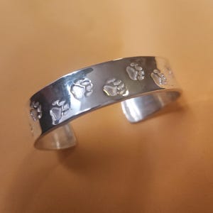 May include: A silver bracelet with paw print designs. The bracelet is open and has a smooth, polished finish.