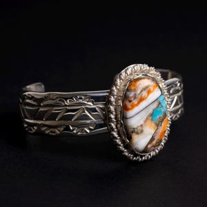 May include: A silver cuff bracelet with a large oval-shaped stone set in the center. The stone is a mix of white, orange, and turquoise colors. The bracelet has a decorative design around the edge.