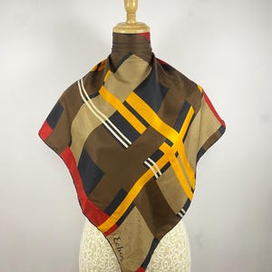 Peut inclure: Un foulard carré en soie présentant un motif géométrique de blocs marron, noirs, jaunes et rouges avec de fines rayures blanches. Le foulard est drapé autour d'un cou de mannequin et affiche le nom de marque Echo imprimé en bas en écriture noire.
