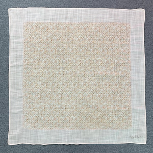 May include: A square white handkerchief with a floral pattern in the center. The floral pattern is a mix of small brown and white flowers. The border is white with a stitched edge. The handkerchief is made of a lightweight fabric.