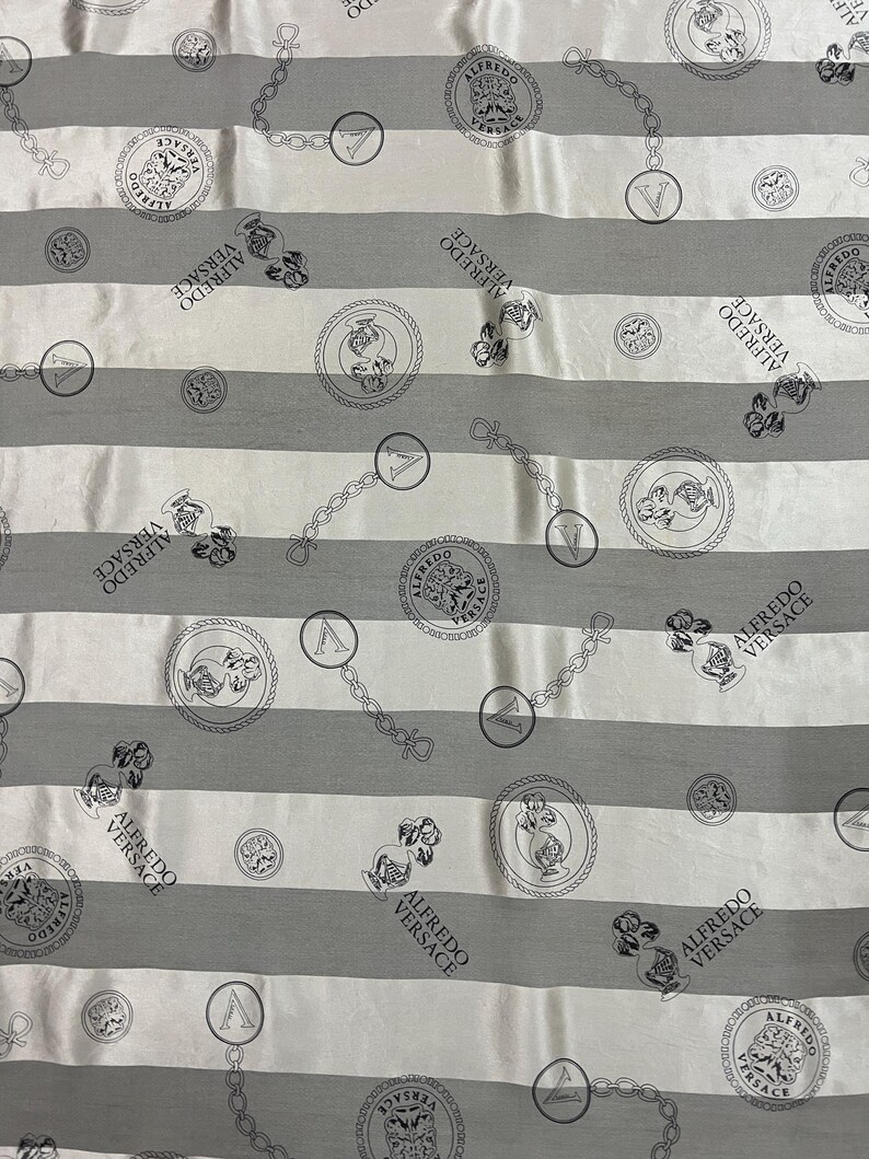 May include: A fabric with a repeating pattern of gray and white horizontal stripes. The design includes circular medallions, chain links, and the text "ALFREDO VERSACE" in a repeating pattern.