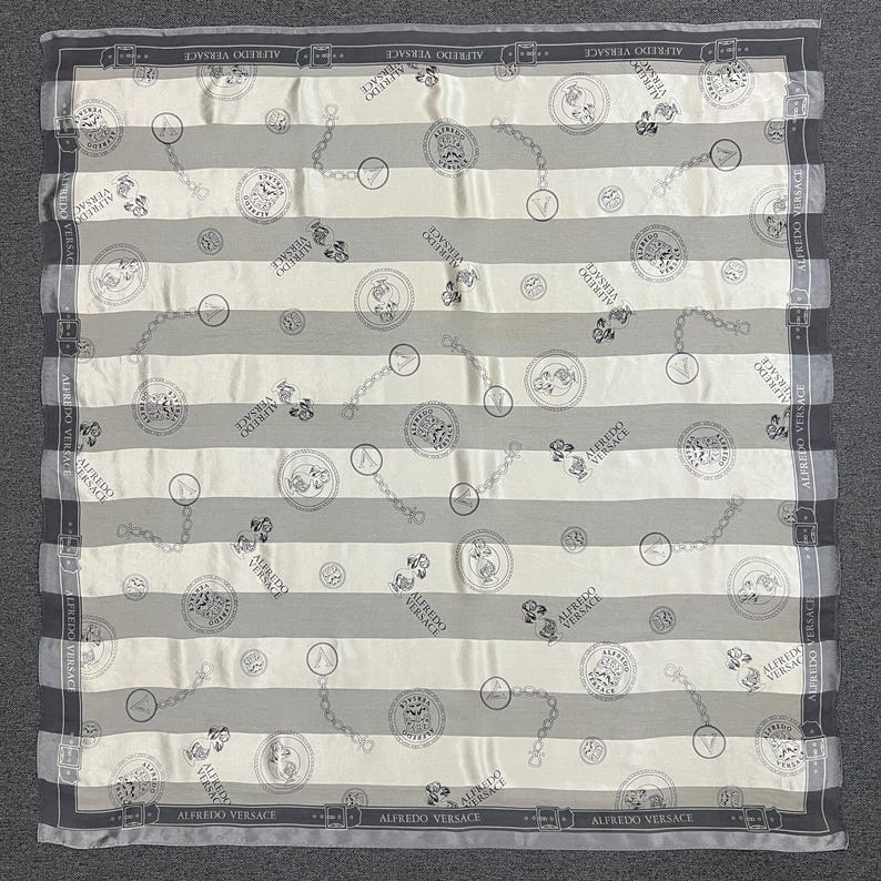 May include: A square silk scarf with a striped pattern in gray and white. The scarf features repeating circular medallions, chain links, and the text "ALFREDO VERSACE" along the borders. The scarf is a luxury fashion accessory.