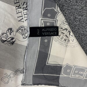 May include: A silk scarf with a repeating pattern in shades of gray and white. The scarf features the text "ALFREDO VERSACE" and a label that reads "ALFREDO VERSACE ITALY". The scarf is draped over a gray fabric.