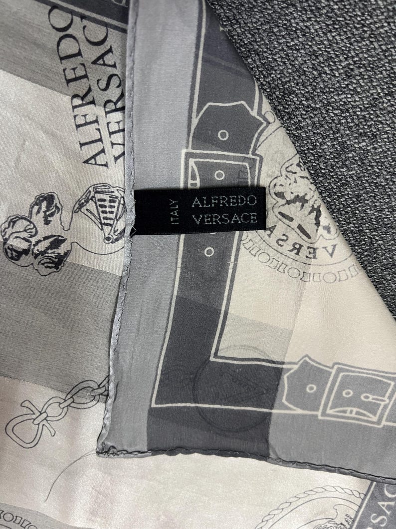 May include: A silk scarf with a repeating pattern in shades of gray and white. The scarf features the text "ALFREDO VERSACE" and a label that reads "ALFREDO VERSACE ITALY". The scarf is draped over a gray fabric.