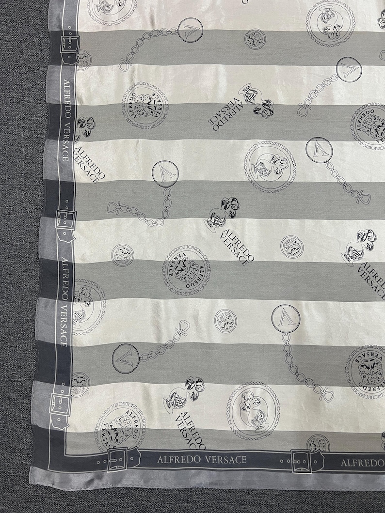 May include: A scarf with a striped pattern in white, light gray, and dark gray. The scarf features repeating circular medallions, chain links, and the text "ALFREDO VERSACE". The edges are bordered with a dark gray design.