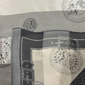 May include: A gray and white silk scarf with a repeating pattern of medallions, chains, and belt buckle designs. The scarf features the text "ALFREDO TOLEDO" in a circular pattern around a lion's head.