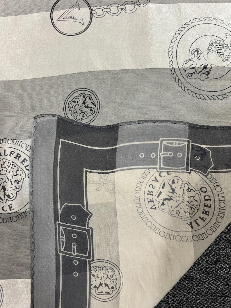 May include: A gray and white silk scarf with a repeating pattern of medallions, chains, and belt buckle designs. The scarf features the text "ALFREDO TOLEDO" in a circular pattern around a lion's head.