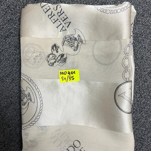 May include: A folded, off-white silk scarf with a repeating pattern of black medallions, chains, and text that reads "ALFRED VERS". A yellow sticker with the text "MO401 34/35" is attached to the scarf.