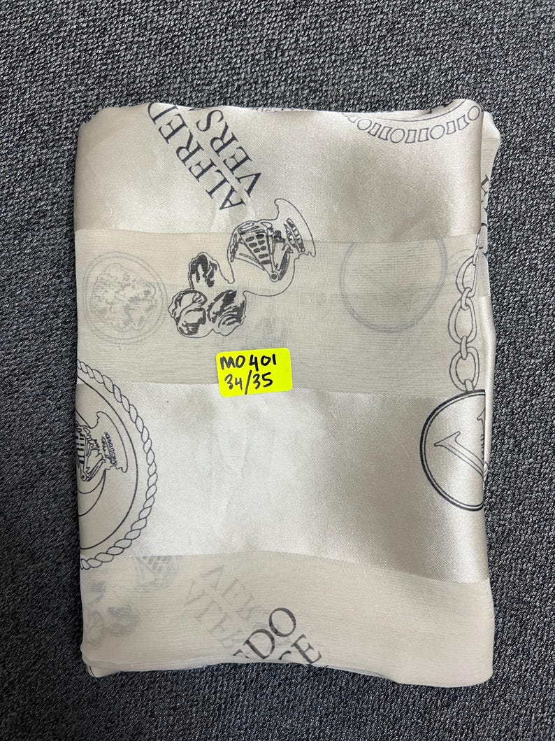 May include: A folded, off-white silk scarf with a repeating pattern of black medallions, chains, and text that reads "ALFRED VERS". A yellow sticker with the text "MO401 34/35" is attached to the scarf.
