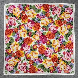 May include: Square floral handkerchief featuring a vibrant pattern of roses, dahlias, and other flowers in shades of pink, yellow, orange, and red. The flowers are set against a white background with green leaves. The handkerchief has a white border and a small "KENZO" logo.