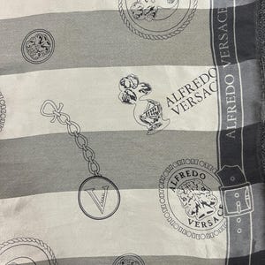 May include: A silk scarf with a striped pattern in shades of gray and white. The scarf features repeating circular medallions, chain links, and the text "ALFREDO VERSACE" in a decorative font. The design is detailed and elegant.