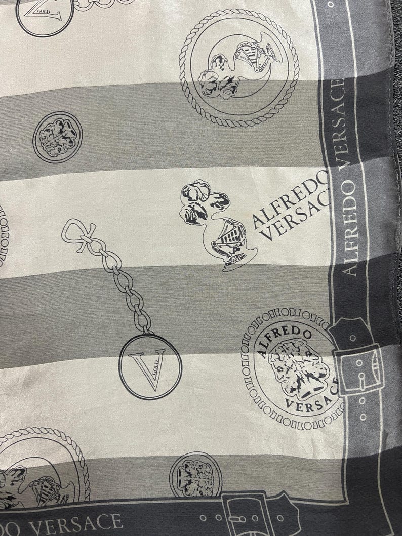 May include: A silk scarf with a striped pattern in shades of gray and white. The scarf features repeating circular medallions, chain links, and the text "ALFREDO VERSACE" in a decorative font. The design is detailed and elegant.