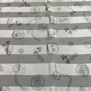 May include: White and gray striped fabric with repeating circular and chain link patterns. The circles contain illustrations and the text "ALFREDO VERSACE". The fabric has a luxurious sheen.