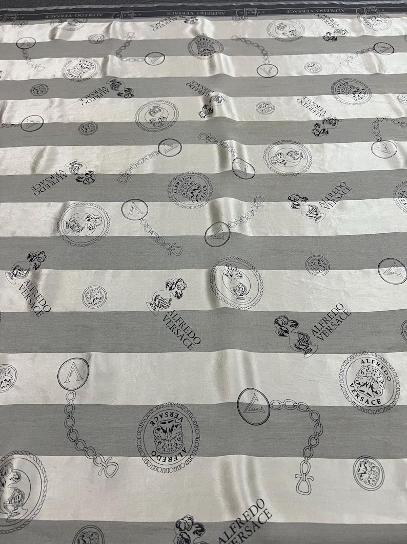 May include: White and gray striped fabric with repeating circular and chain link patterns. The circles contain illustrations and the text "ALFREDO VERSACE". The fabric has a luxurious sheen.