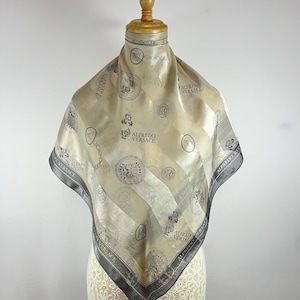 May include: A square silk scarf in cream, gray, and black, draped on a mannequin. The scarf features the text "ALFREDO VERSACE" and a repeating pattern of medallions, chains, and figures. The border is a dark gray.