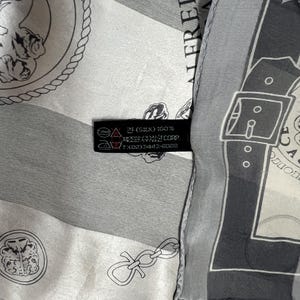 May include: A silk scarf with a repeating pattern of medallions, chains, and geometric shapes in shades of gray and white. The scarf has a label with text including "SILK 100%" and the brand name "ALFREDO".