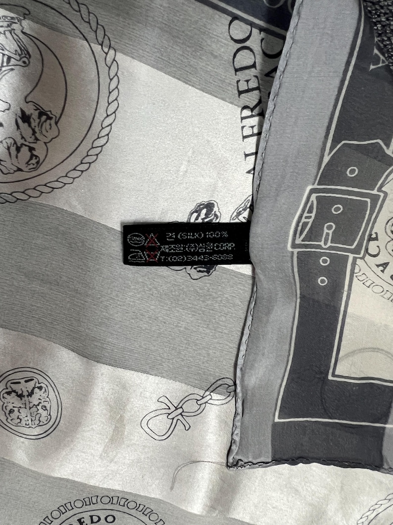 May include: A silk scarf with a repeating pattern of medallions, chains, and geometric shapes in shades of gray and white. The scarf has a label with text including "SILK 100%" and the brand name "ALFREDO".