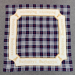 May include: Square scarf with a classic plaid pattern in navy blue, red, and white. The scarf features a cream-colored border with the text "Burberrys of London" in gold lettering, and a gold logo.