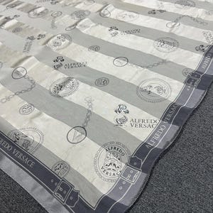 May include: A silk scarf with a striped pattern in shades of gray and white. The scarf features repeating circular medallions, chain links, and the text "ALFREDO VERSACE" along the border. The design is elegant and suitable for fashion accessories.