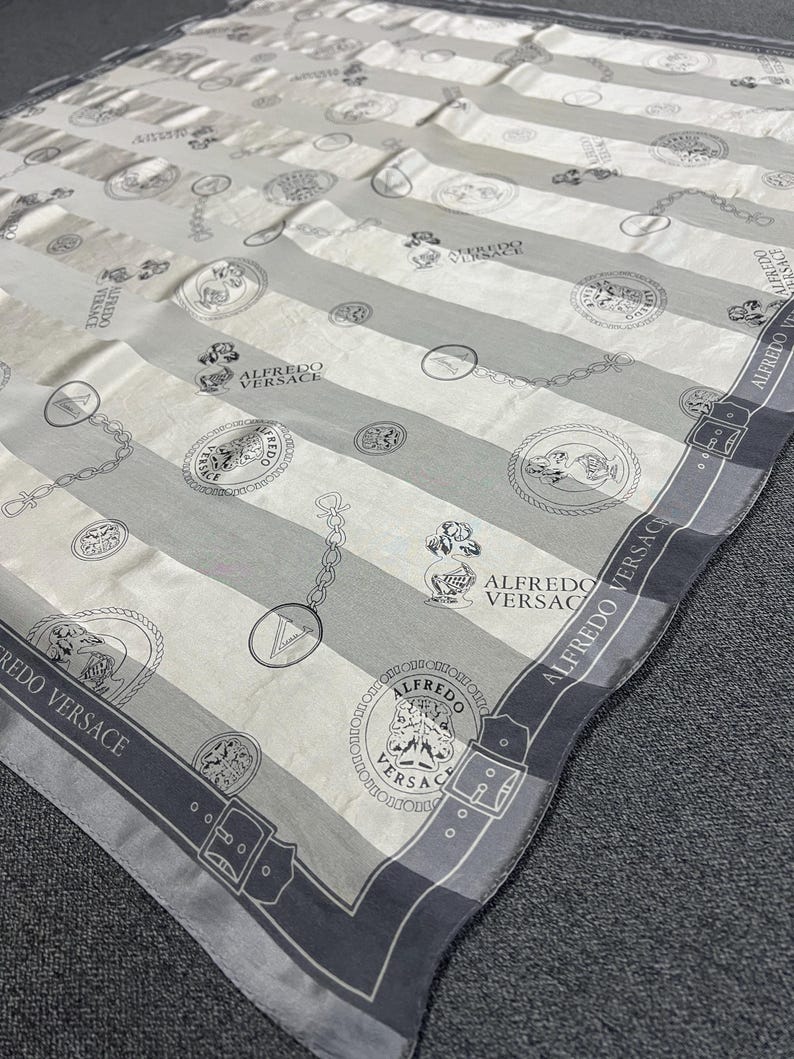 May include: A silk scarf with a striped pattern in shades of gray and white. The scarf features repeating circular medallions, chain links, and the text "ALFREDO VERSACE" along the border. The design is elegant and suitable for fashion accessories.
