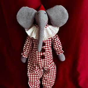 May include: A gray stuffed elephant wearing a red and white checkered jumpsuit with a white ruffle collar and a red hat. The elephant is standing on a red background.