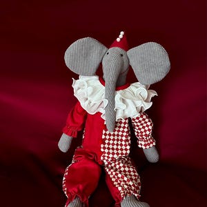May include: A gray stuffed elephant wearing a red and white checkered jumpsuit with a white ruffle collar and a red hat with a white pearl on top. The elephant is sitting on a red satin surface.