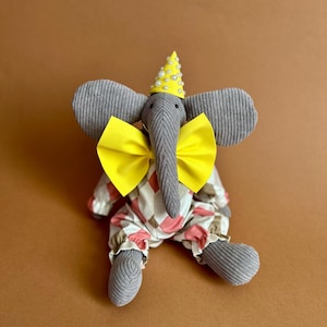 May include: A gray stuffed elephant wearing a yellow and white party hat, a yellow bow tie, and a pink and white checkered jumpsuit.